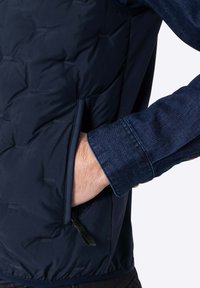 Marineblaues isoliertes Westchen mit strukturierter Oberfläche, Seitenfach und verstellbarem Saum, getragen über einem Jeanshemd mit umgeschlagenem Ärmel.