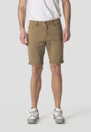 Hombre con pantalones cortos marrones enrollados hasta la rodilla, camiseta blanca y zapatillas blancas, de pie sobre un fondo sencillo de color gris claro.