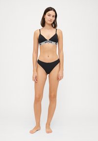 Μαύρο σετ μπικίνι που περιλαμβάνει μια μπράλετ με τρίγωνο σχήμα και λεπτές τιράντες, καθώς και ελαστική γκρι ζώνη με επωνυμία. Συμπληρώνεται από χαμηλοκάβαλα μπικίνι σλιπ. Απαλή ύφανση.