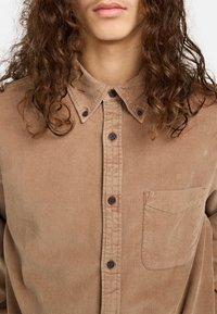 Braunes Cordhemd mit button-down Kragen, ausgestattet mit einer Brusttasche und braunen Knöpfen. Textil mit strukturiertem Stoff und senkrechten Rippungen.