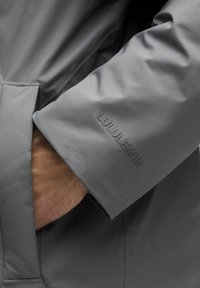 Käsi on sisestatud halli Lululemoni jope taskusse, näidates brändi nime, mis on reljeefselt väljatrükitud varruka ääres.