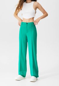 Femme portant un pantalon droit taille haute turquoise, un crop top blanc et des baskets blanches, debout avec les mains près de la ceinture.
