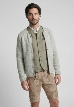 Mann trägt traditionelle bayerische Kleidung, darunter bestickte Lederhose, geknöpfte Weste, weißes Hemd und hellgraue gestrickte Jacke.