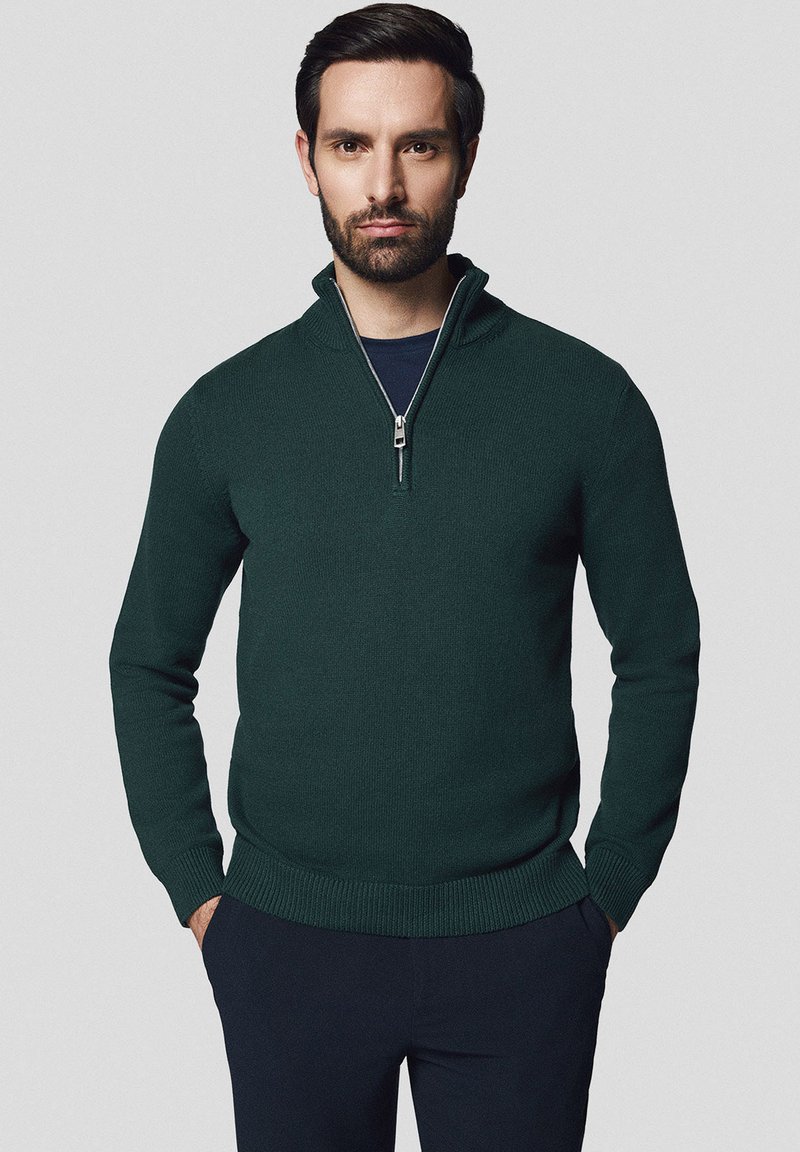 Ciemnozielony sweter z zamkiem na szyi, wykonany z dzianiny, z długimi rękawami i ściągaczami na mankietach. Noszony z granatową koszulą pod spodem.