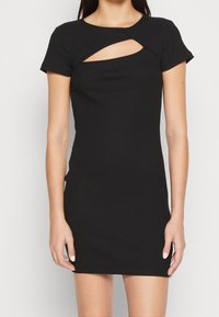 Femme portant une robe noire cintrée à manches courtes en côte avec une découpe diagonale au niveau de la poitrine, se tenant devant un fond clair.