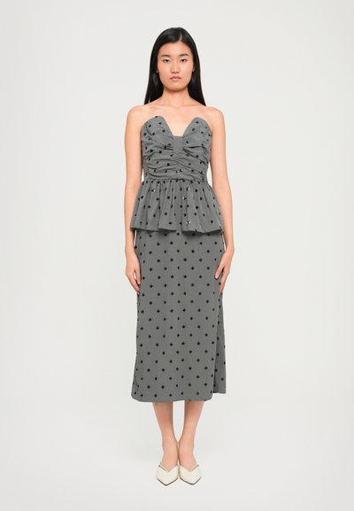 Robe péplum grise avec encolure en cœur, corsage froncé et motifs en losange noirs, associée à des chaussures blanches à bout pointu.