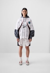 White oversized hoodie with gray sleeves; features black and red accents and various signatures. Paired with black shorts and red-tinged footwear.