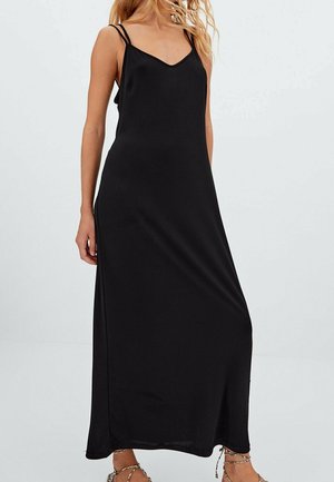 Woman wearing a sleeveless black maxi dress with double thin straps and braided ankle-strap sandals, standing against a plain background.