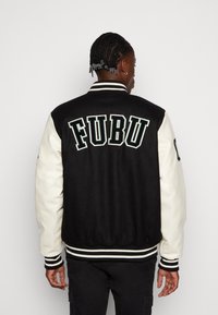 Black and cream varsity jacket with "FUBU" embroidered on the back, striped ribbed cuffs, and waist. Smooth fabric texture. Regular fit.