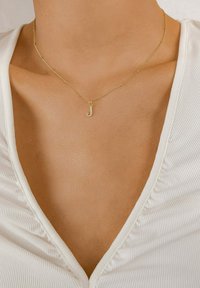Collier en or avec un pendentif en forme de "J" orné de petites pierres transparentes, reposant sur une clavicule nue, le haut texturé blanc étant visible.