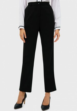 Frau trägt schwarze, gerade geschnittene Hose, weißes Langarmshirt mit schwarzem Besatz an den Bündchen und schwarze High Heels.