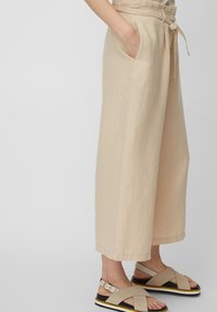 Beżowe lniane spodnie w krótkim kroju z szerokimi nogawkami, z elastycznym pasem i bocznymi kieszeniami. W zestawieniu z jasnymi neutralnymi sandałami z krzyżującymi się paskami.