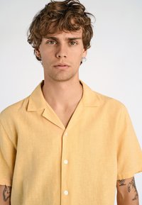 Camicetta a maniche corte, con bottoni, in tessuto di lino giallo chiaro. Presenta un colletto con angolo e bottoni bianchi, con una vestibilità ampia.