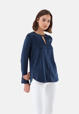 Marineblaue Seidenbluse mit einem Schlüsselloch-Ausschnitt, langen Ärmeln und Seitenschlitzen, kombiniert mit weißen Hosen für den Kontrast.
