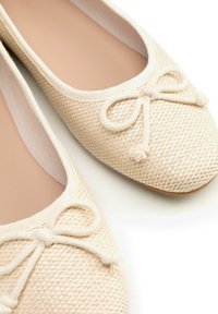 Beige geweven ballerina's met afgeronde neuzen en decoratieve strikjes bovenop, getoond op een witte achtergrond.