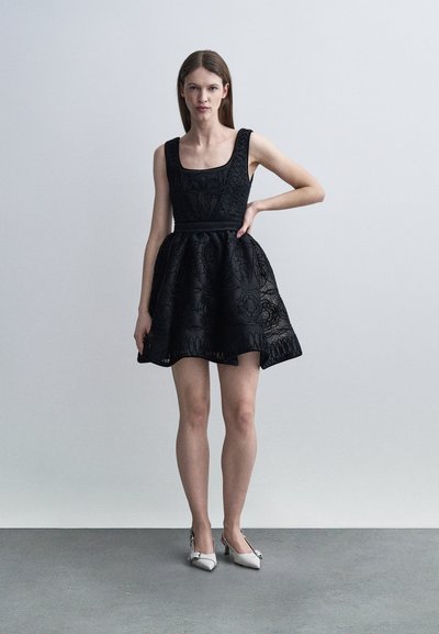 Γυναίκα που στέκεται, φορώντας κοντό μαύρο φόρεμα χωρίς μανίκια με υφή και άσπρα παπούτσια με χαμηλό τακούνι, μπροστά σε απλό φωτεινό φόντο.