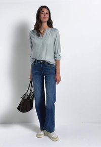 Blouse longue à manches vert clair avec un col à boutons, associée à un jean à jambe large bleu foncé et un sac en maille marron. Chaussures blanches aux pieds.
