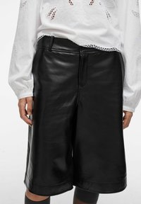 Personne portant un short en cuir noir jusqu'aux genoux et un haut à manches longues blanc avec des broderies en dentelle, les mains détendues sur les côtés.