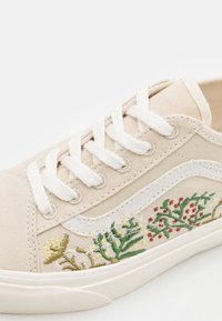 Baskets en toile beige avec lacets et semelle blancs, ornées de motifs floraux brodés en vert, rouge, jaune et blanc sur le panneau latéral.