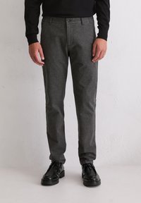 Mann in schmal geschnittenen, grauen karierten Hosen, schwarzem Langarmhemd und schwarzen, geschnürten Lederschuhen, der an einer weißen Wand steht.