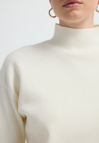 Kremowy sweter z wysokim kołnierzem, wykonany z miękkiego materiału, o luźnym kroju i prążkowanej teksturze, w zestawie z srebrnymi kolczykami w kształcie kół.