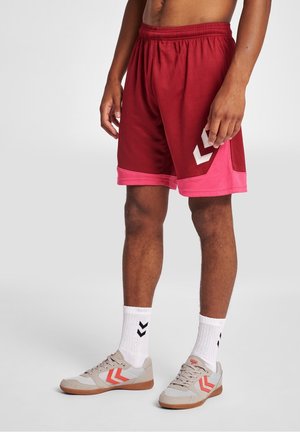 Man wearing red and pink athletic shorts, white socks with black chevrons, and beige sneakers with orange chevron designs, standing on a white floor.
