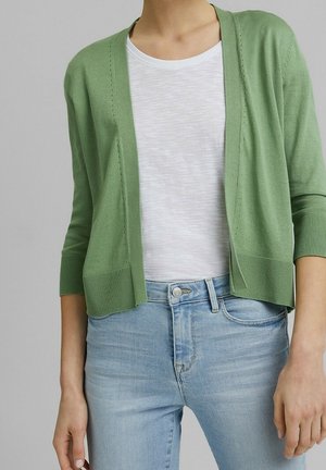 Osoba w jasnozielonym kardiganie z otwartym przodem, na białej koszulce i jasnoniebieskich dżinsach, stojąca na tle jednolitego, szarego tła.