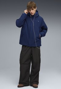 Tumši zilas krāsas jaka ar kapuci ar teksturētu apdari, priekšējām kabatām, uzvilkta virs melnajām biksēm ar kabatām. Redzamas austiņas, tumšs fons.