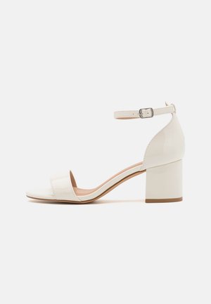 Hvid patentlæder sandal med hæl, ankelrem, blokhæl og åben tå mod en ensfarvet baggrund.