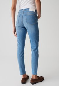 Jean bleu clair à coupe slim, avec cinq poches, une fermeture éclair et des détails de couture subtils. Porté avec des mocassins marron.