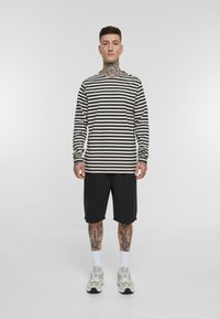 Chemise à rayures noir et blanc à manches longues, shorts noirs, chaussettes blanches et baskets claires. Tatouages visibles sur les bras et les jambes.