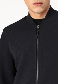 Tume bomber-jakk, mis on valmistatud tekstureeritud kangast, kõrge kaelusega, ees lukuga ja peene mustriga läbi kogu jaku.