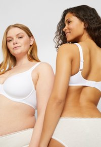 Twee vrouwen in witte bh's en crèmekleurig ondergoed poseren vol zelfvertrouwen, de een kijkt recht vooruit en de ander draait haar rug naar de camera.