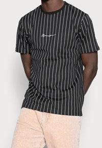 Černé tričko s krátkým rukávem s vertikálními bílými pruhy a malým bílým logem na hrudi. Klasický kulatý výstřih, vyrobeno z džersejového materiálu.