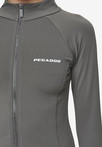 Pilkas zip-up sportinis džemperis su ilgomis rankovėmis, turintis aukštą apykaklę ir mažą baltą "PEGADOR" logotipą krūtinėje. Lygus, tekstūruotas audinys.