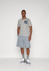 Pilkas medvilninis marškinėliai su tamsiai mėlynu Niujorko "Yankees" logotipu, derinami su šviesiai pilkomis �šortais. Nešiojami su pilkais Nike kojiniais ir juodai baltais sportbačiais.