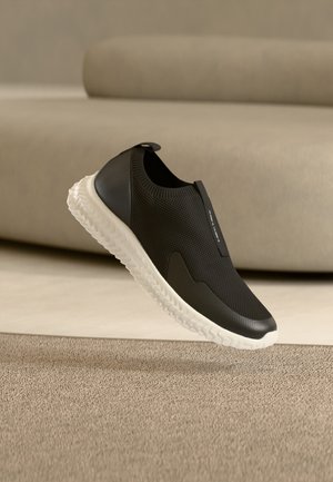 Sneaker nera slip-on con suola bianca strutturata che fluttua sopra un tappeto beige, su uno sfondo imbottito tono neutro.