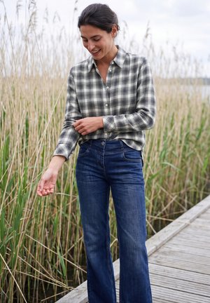 Femme portant une chemise à carreaux et un jean bleu, debout sur un quai en bois à côté de hautes herbes, souriante et regardant son bras.