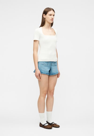 Γυναίκα που στέκεται στραμμένη προς τα δεξιά, φορώντας λευκή μπλούζα με τετράγωνη λαιμόκοψη, τζιν σορτς, λευκές κάλτσες και φλατ παπούτσια με λεοπάρ μοτίβο, σε απλό φωτεινό φόντο.