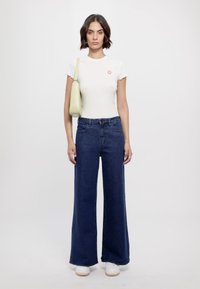 Top alb cu mânecă scurtă, cu margini ciupite, blugi largi albaștri și teniși albi, accessorizați cu o geantă verde de umăr.