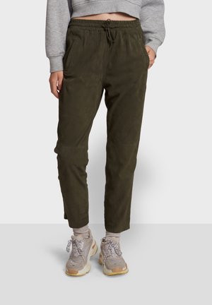 Person trägt olivgrüne Jogginghosen mit Kordelzug und Seitentaschen, hellgraue Socken, klobige beige Sneaker und ein graues, kurz geschnittenes Sweatshirt.