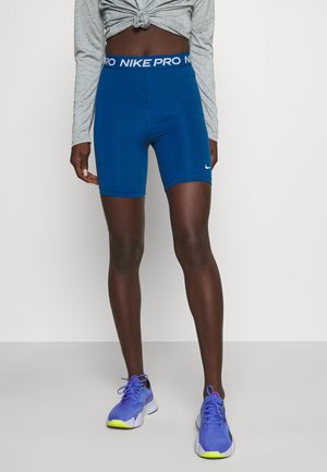 Atletisk person iført blå Nike Pro-shorts, grå langærmet top knyttet i taljen og blå løbesko med gule såler, stående på gråt gulv.
