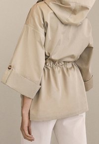 Beżowa kurtka z kapturem, z taliowanym pasem, szerokimi rękawami i ozdobnymi guzikami. Wykonana z błyszczącego materiału o gładkiej fakturze. Widok z tyłu.