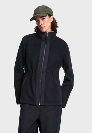 Femme portant une veste noire zippée à col montant, un pantalon noir et une casquette vert olive sur un fond gris clair uni.
