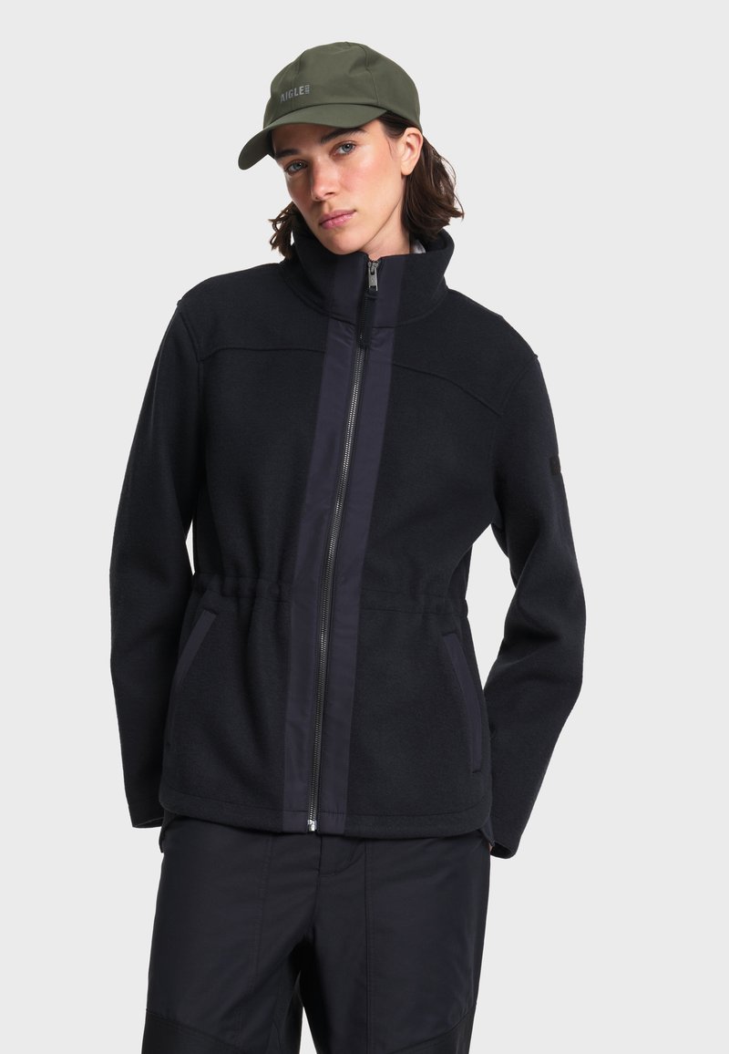 Femme portant une veste noire zippée à col montant, un pantalon noir et une casquette vert olive sur un fond gris clair uni.
