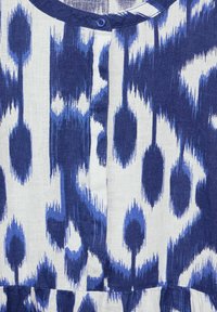 Blau-weiß gemusterter Stoff mit einem Ikat-Design; hat einen runden Ausschnitt und einen einzelnen blauen Knopf in der Mitte. Weiche Textur.