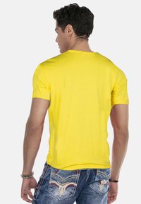 Gelbes T-Shirt mit kurzen Ärmeln aus Baumwolle, das ein schlichtes Design aufweist. Kombiniert mit blauen Jeans aus Denim, die detaillierte Nähte an den Taschen zeigen.