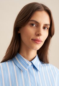 Camicia collare azzurra con strisce verticali bianche e blu scuro alternate, dotata di chiusura a bottone al collo e texture liscia.