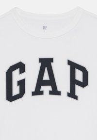 Biała bawełniana koszulka z odważnym czarnym napisem "GAP" w stylu uniwersyteckim. Koszulka ma okrągły dekolt i miękką teksturę.