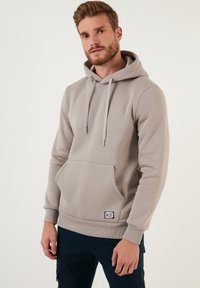 Sudadera gris claro hecha de una tela suave, con un bolsillo canguro frontal, cordones y una pequeña etiqueta de marca.